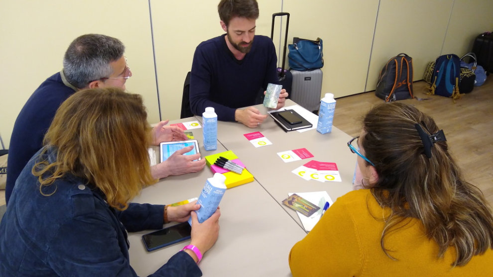 Photograph of a group of participants. One of them is holding a Dixit card and explaining something to the others. One of the participants is taking notes on an iPad.